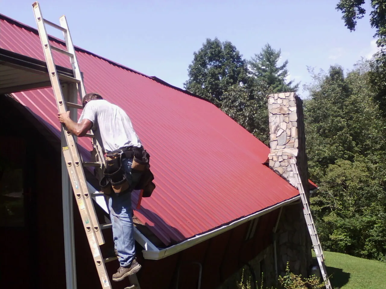 Roofer on a ladder beside a red metal roof and stone chimney — Soffit & Fascia Repair by Roofing Brothers in Broadview Heights